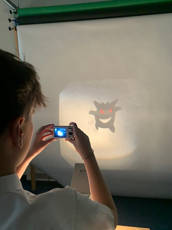 Photograph showing a person using a camera to capture a shadow puppet of a character with spiky hair and glowing red eyes projected onto a white screen. The scene highlights creative use of lighting and shadow to create a striking visual effect with a green backdrop partially visible above the screen.