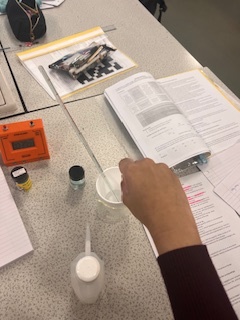 Photograph showing a person conducting a science experiment at a lab table with an open textbook, small containers, and a pipette. The scene highlights practical learning with labelled equipment and study materials arranged for reference and measuremen