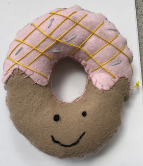 Photograph of a handmade felt donut plush toy featuring a brown base with a smiling face and pink frosting decorated with yellow and light blue stitched lines. The plush has visible hand stitching around edges and a hole in the centre, giving it a soft, whimsical appearance.