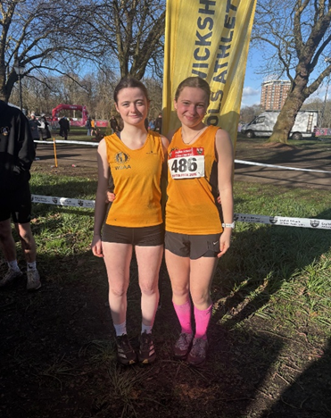 Photograph of two female athletes standing side by side outdoors, wearing matching orange sleeveless tops and black shorts, with one athlete wearing a race bib numbered 486. They are positioned in front of a yellow banner with partially visible text related to athletics