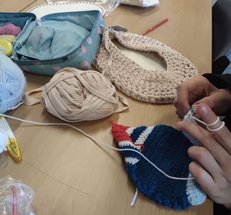 Photograph showing a person crocheting a blue, red, and white piece with yarn and crochet hook on a wooden table. Surrounding items include a beige crocheted bag, a ball of beige yarn, a blue yarn ball, and a floral-patterned open bag with knitting supplies.