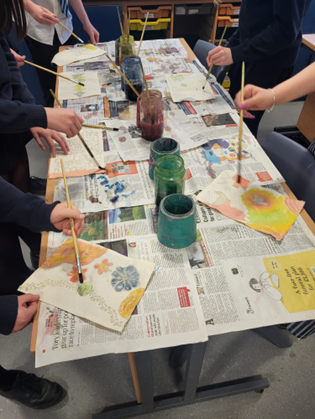 Photograph showing multiple people painting floral and abstract designs on paper using watercolours at a table covered with newspapers. Several jars with different coloured paints are placed in the centre, with brushes held by participants actively working on their artwork.