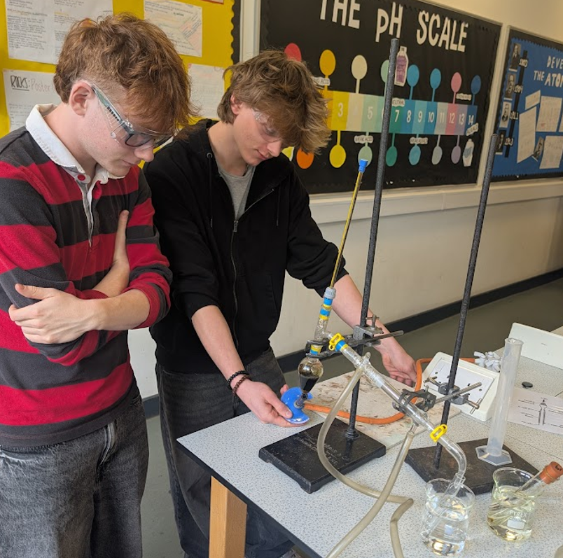 Photograph of two students conducting a chemistry experiment in a classroom, with a pH scale poster and science-related displays on the wall behind them. The setup includes glassware, tubing, and a stand with clamps, indicating a focus on measuring