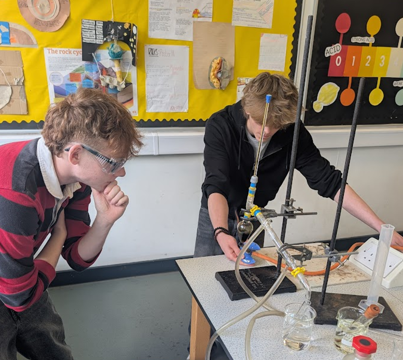 Photograph of two students conducting a chemistry experiment in a classroom with science-themed posters on a yellow bulletin board. Equipment includes glassware, tubing, and clamps, with one student adjusting apparatus