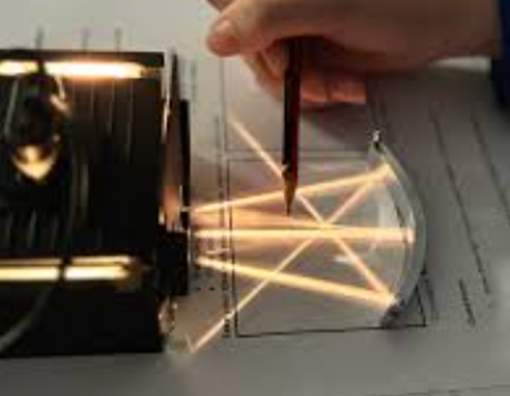 Photograph showing a hand holding a pencil to trace light rays passing through a curved transparent lens on a diagram. The setup demonstrates light refraction and reflection patterns with visible light beams and labelled grid lines on the paper beneath.