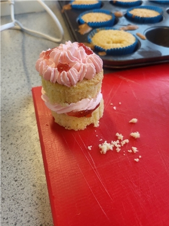 Photograph showing a cupcake with pink frosting and red jam filling placed on a red cutting board, with crumbs scattered nearby. In background, a baking tray holds several un iced cupcakes in blue paper cases on a speckled countertop.