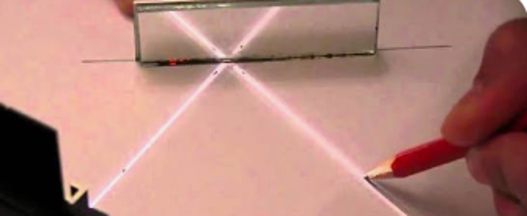 Photograph showing a laser beam being split by a beam splitter, creating two intersecting light paths on a white surface. A hand holding a red pencil is marking the point where the beams cross, illustrating an experiment setup for studying light interference or reflectio
