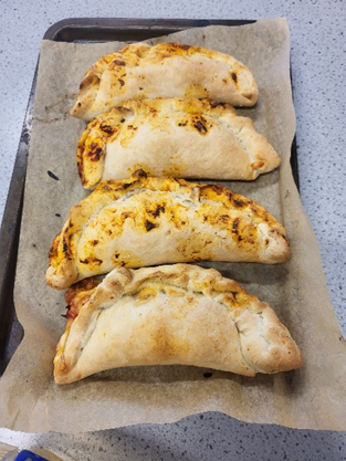 Photograph of four baked calzones arranged in a row on parchment paper atop a baking tray. Calzones vary slightly in size and browning, with golden crust