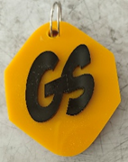 Photograph of a yellow octagonal keychain with black raised letters "GS" placed on a  white surface. 