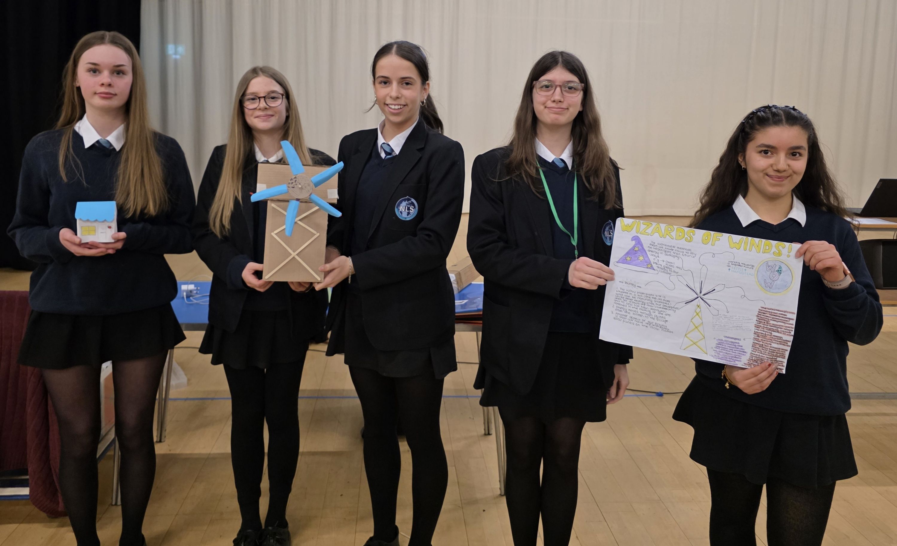 Photograph of five students in uniform, each holding different objects including a small box, a windmill model, and a poster with drawings and text