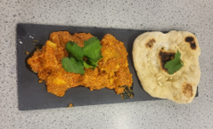 Photograph of a serving of Indian-style curry with naan bread on a rectangular black slate plate. Curry is orange with visible chunks, garnished with fresh green coriander leaves, and naan has a small coriander leaf and toasted spots.