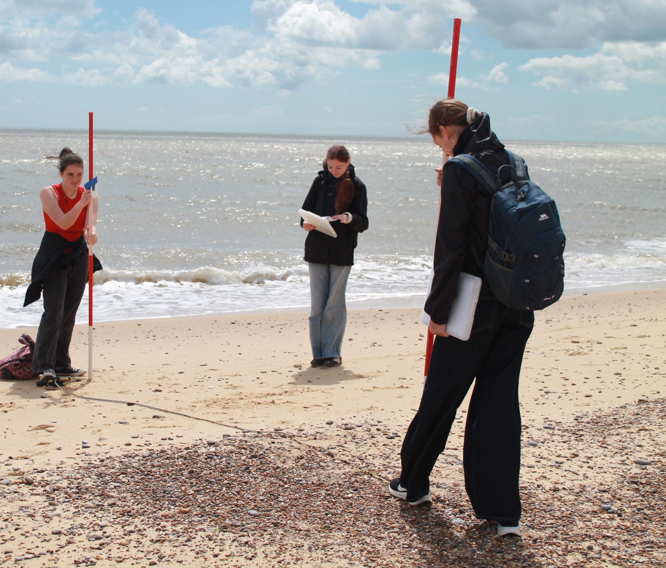 Photograph showing three people conducting a beach survey or measurement activity, using red poles and a measuring tape stretched across sand and pebbles near the shoreline. Two individuals hold poles at each end of the tape while the third person reads notes, with cloudy sky and calm sea in background.