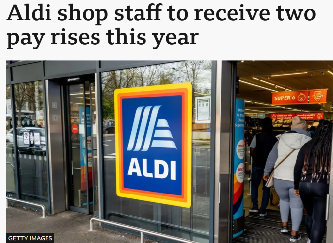 Photograph showing entrance of Aldi store with prominent blue and yellow Aldi logo on glass door and several customers entering. Context highlights news about Aldi shop staff receiving two pay rises this year, indicating positive employee wage developments.