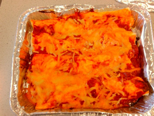 a baked dish in a rectangular aluminium foil container, featuring layers of pasta covered with melted orange cheese and red tomato sauce. The dish appears freshly cooked with cheese evenly spread and slightly browned in some areas.