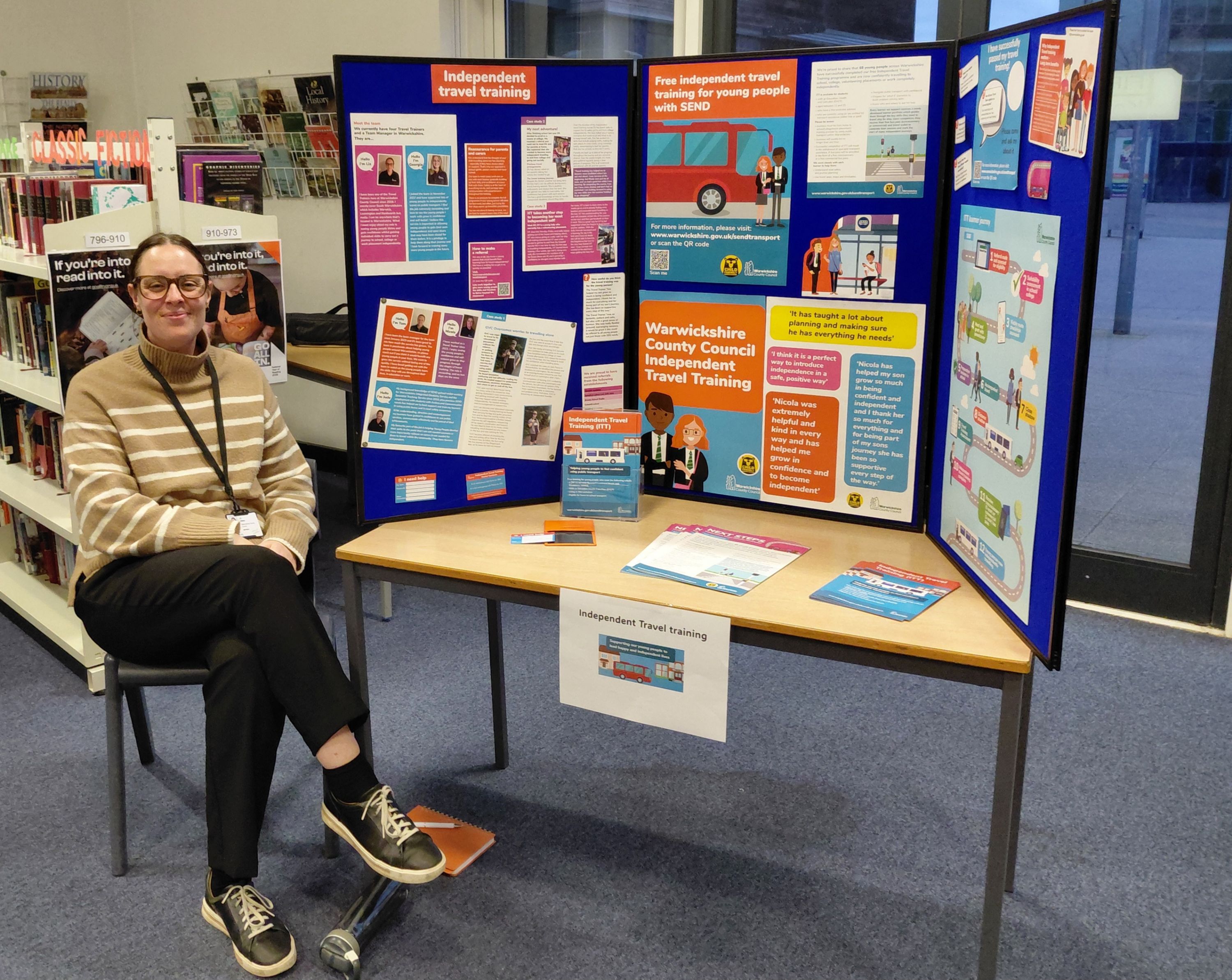 Photograph of an information display board set up on a table promoting Warwickshire County Council Independent Travel Training, featuring colourful panels with text, images, and illustrations about travel skills and support for young people. A person sits beside the table in a library setting, with brochures and flyers arranged on the table, highlighting training benefits and contact information.