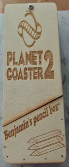 Photograph of a wooden pencil box engraved with "Planet Coaster 2" and "Benjamin's pencil box," featuring a roller coaster graphic and three pencil illustration