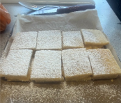 Photograph showing nine square pieces of mille fuille  dusted with powdered sugar on baking paper-lined tray. Knife and orange peeler are visible on countertop beside tray, indicating freshly baked and prepared treats.