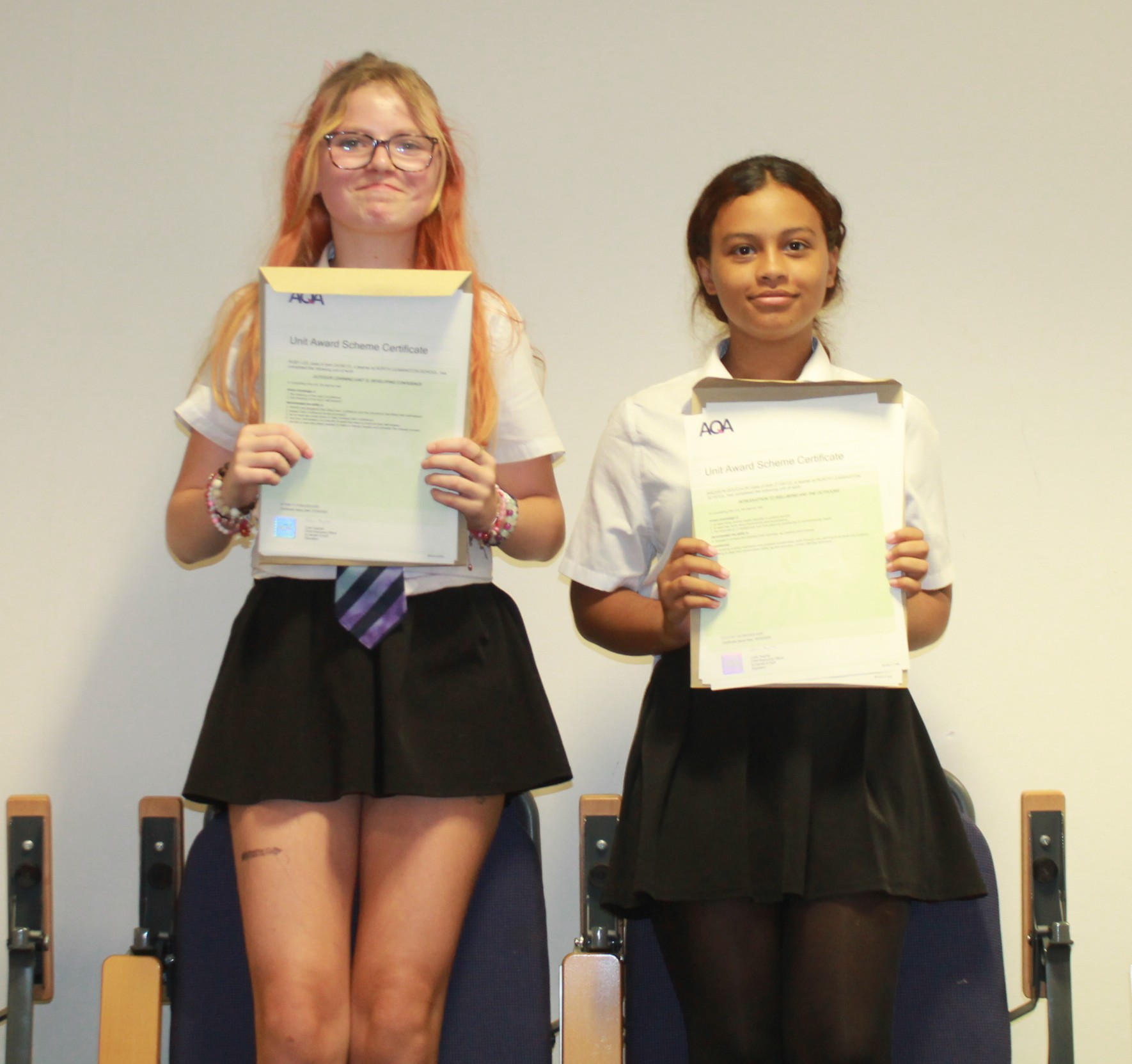 Photograph of two girls standing in a row holding certificates, likely celebrating an academic or extracurricular achievement. They wear white shirts and black trousers, with a plain background and chairs partially visible behind them