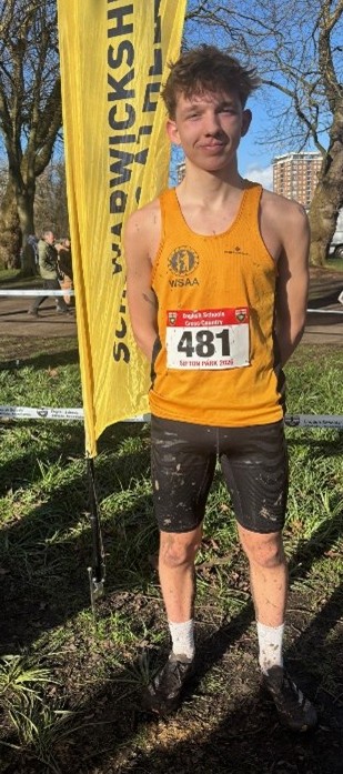Photograph of a male runner standing outdoors on muddy grass near a yellow vertical banner with "SOUTHWICKSHIRE" text. The runner wears a yellow singlet with a race number 481 and black shorts