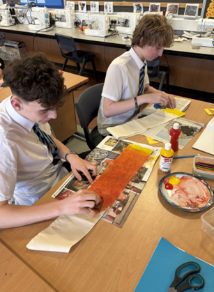Photograph of two students in a classroom engaged in fabric painting activities, with sewing machines and instructional posters visible in the background. One student is applying orange paint on fabric, while the other works with yellow paint, with two paint bottles, a palette, and scissors on the table.