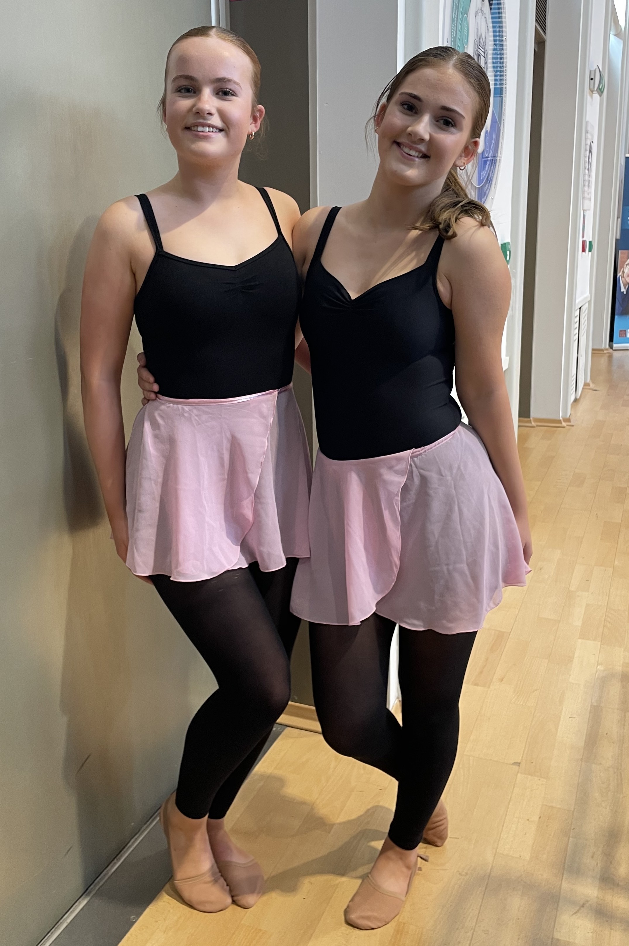 Photograph of two dancers standing side by side in a hall with wooden flooring and a grey wall. Both wear black leotards, pink skirts, black tights, and ballet shoes, smiling at the camera with aa door visible in the background.