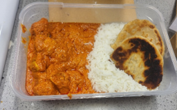 Photograph of a meal in a rectangular plastic container featuring white rice, orange-coloured curry with chunks of chicken, and two pieces of slightly charred flatbread. The meal is neatly divided into three sections, highlighting contrasting textures and colours of the food components.