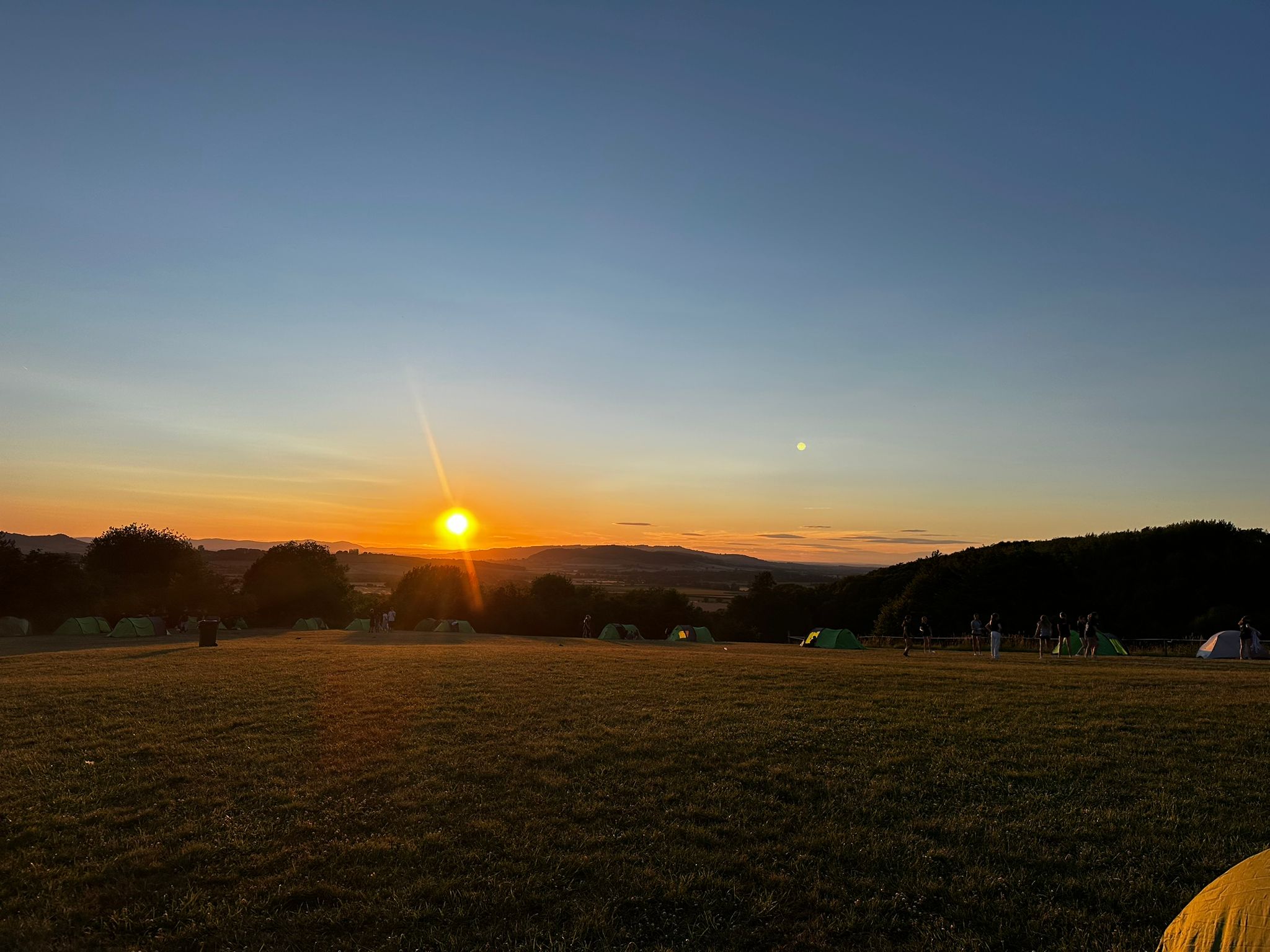 Photograph of a campsite at sunset showing a wide grassy field with multiple tents scattered across it and a clear sky transitioning from orange near horizon to deep blue above. Sun is low near horizon, casting long shadows, with hills and trees in background and a few people visible near tents on right side.