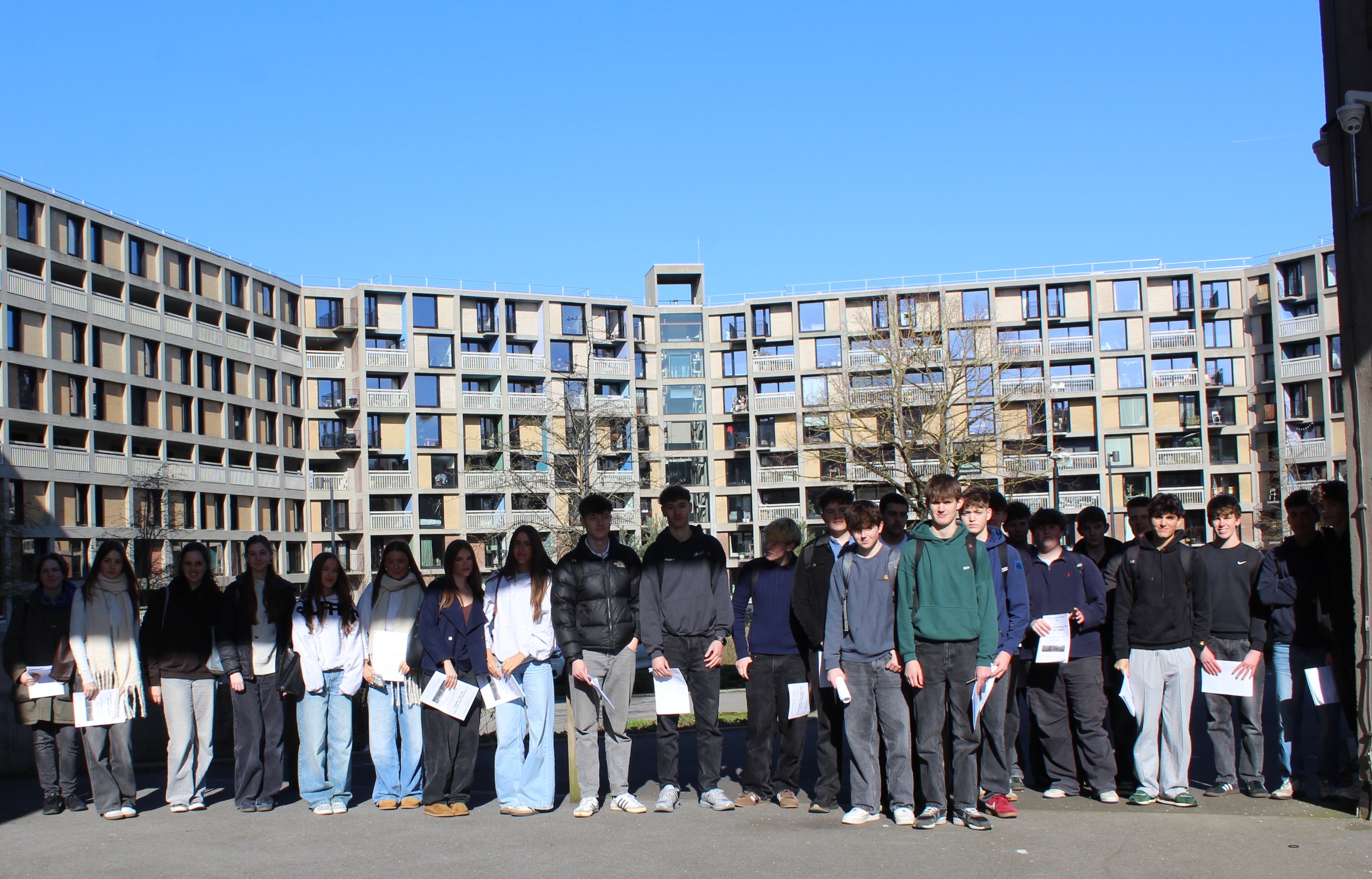 Photograph of a group of students standing in a line outdoors in front of a large, modern, multi-story building with numerous square windows. The group holds papers and wears casual clothing under a clear blue sky