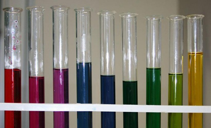 Photograph of nine test tubes in a rack containing liquids of varying colours, ranging from red, purple, blue, green to yellow. The arrangement highlights a spectrum of colours, possibly representing different chemical substances or pH levels.