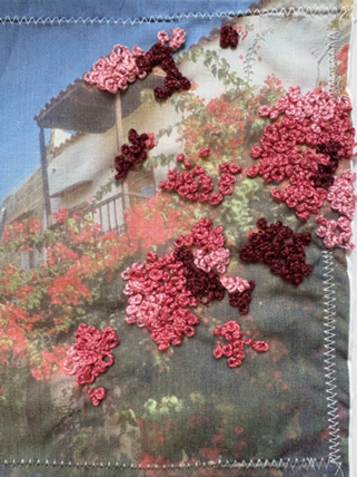 Photograph of fabric featuring a printed outdoor scene with a house and red flowers, enhanced by clusters of pink and dark red embroidered floral patterns. Zigzag stitching borders fabric edges, adding texture and dimension to the mixed-media design.