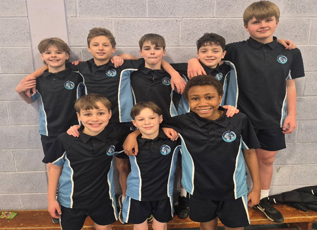 Photograph of eight boys wearing matching black and blue sports uniforms posing indoors against a grey brick wall. Boys are standing and sitting on a wooden bench, with arms around each other, suggesting team camaraderie.