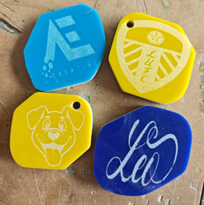 Photograph showing four irregularly shaped plastic key tags in yellow, blue, and dark blue colours arranged on a wooden surface. Each tag features a distinct engraved design, including a cartoon dog face, a stylised "Creative" logo, a cursive script, and a shield emblem with a crown and stripes