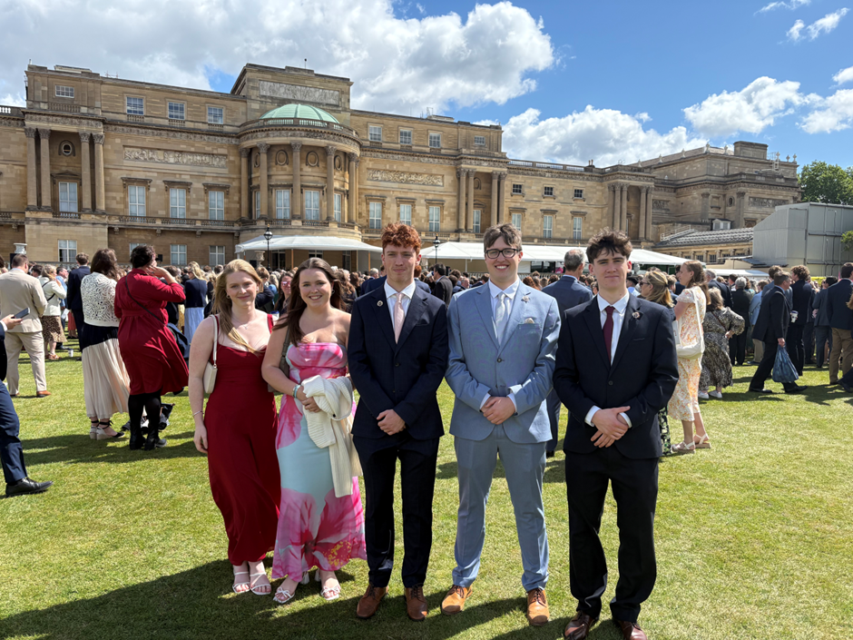 Photograph of a formal outdoor gathering on a lawn in front of a large historic building under a partly cloudy sky. Five young adults dressed in formal attire stand in the foreground, with a crowd of similarly dressed people mingling in the background.