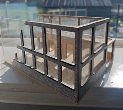         A photograph of a small architectural model depicting a two-storey building with large window openings and a stone-textured exterior. The model highlights structural framework and transparent walls, showcasing an unfinished or skeletal design with clear division between floors.        