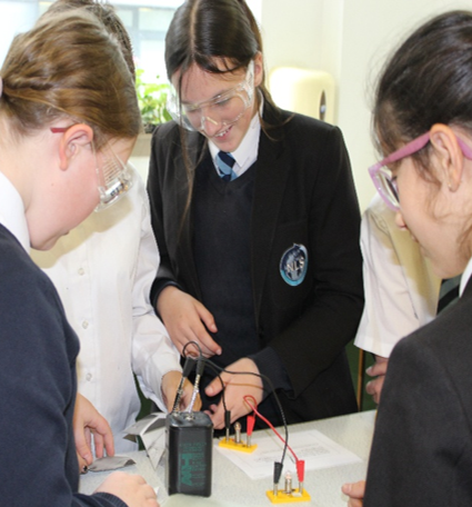 Photograph of four school students wearing safety goggles conducting a science experiment in a classroom. Students are using electrical components and a multimeter on a table, demonstrating hands-on learning and teamwork.