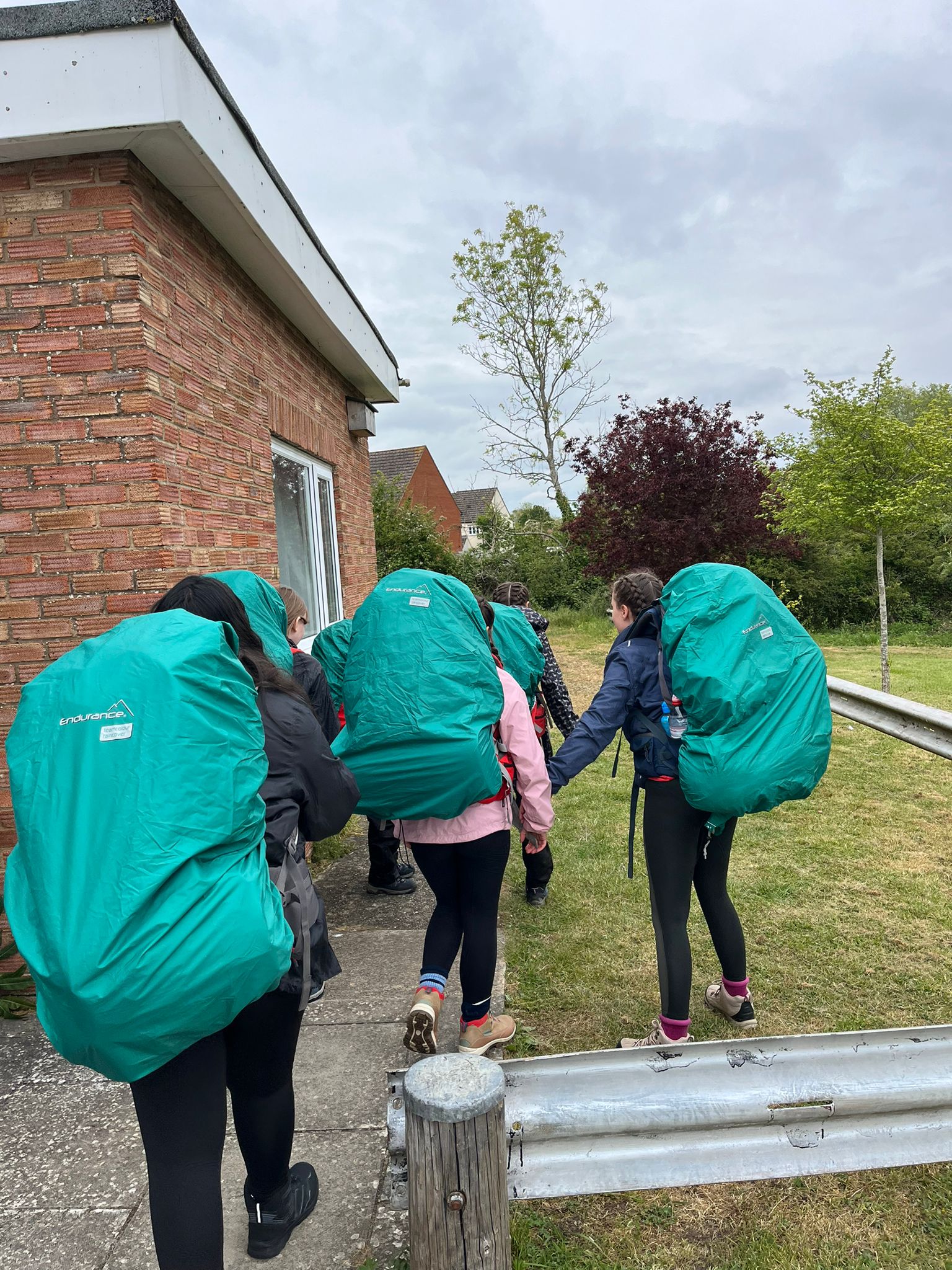 Photograph showing a group of people walking outdoors along a path beside a brick building, each carrying large green backpacks covered with rain covers. The scene captures an outdoor activity or hike, with greenery and trees visible in the background under a cloudy sky.