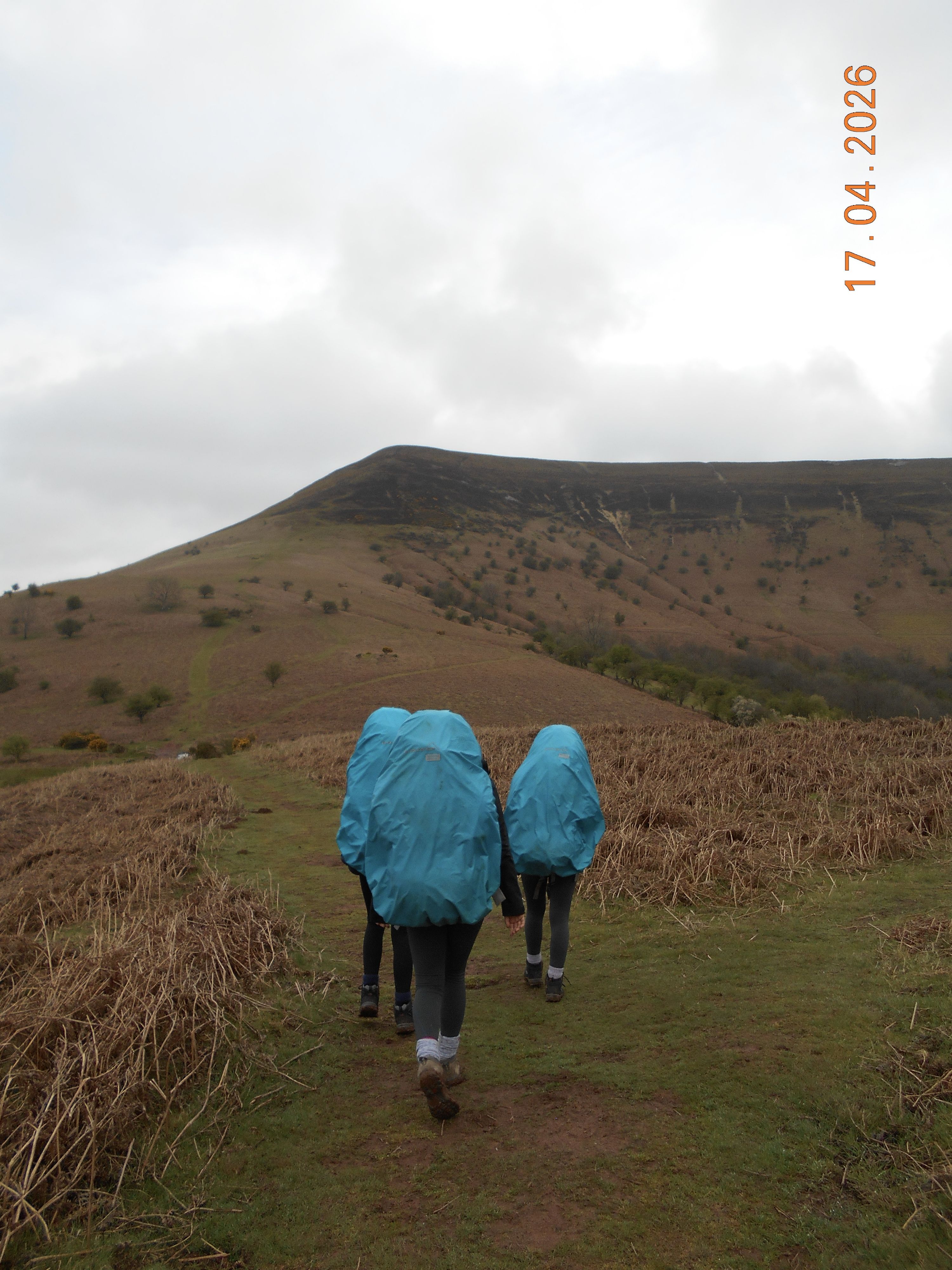 Photograph of three hikers walking on a grassy trail through brown bracken towards a large, rounded hill under a cloudy sky.  hikers wear blue rain covers on their backpacks
