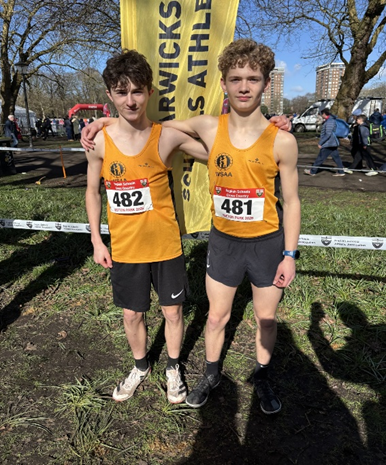 Photograph of two male athletes wearing matching orange singlets and black shorts, standing side by side with arms around each other in an outdoor park setting. Both have race bibs numbered 482 and 481, with a yellow banner behind them displaying "Warwicks Schools Athletics," indicating participation in a cross-country or running event.