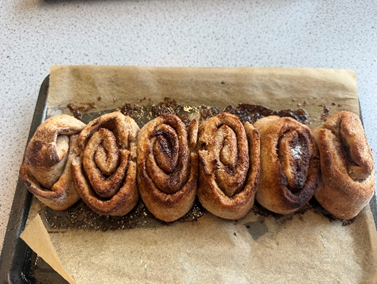 Photograph of a batch of six cinnamon rolls on baking paper atop a baking tray, showing uneven shapes and varying degrees of browning. Rolls are arranged in a row with some cinnamon filling spilled onto the paper, indicating a homemade or rustic baking style.