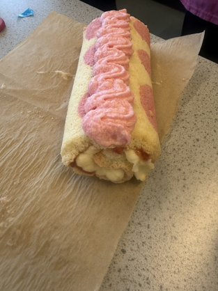 a Swiss roll cake with pink polka dots and a pink cream swirl on top, placed on brown parchment paper on a speckled countertop. The cake is filled with white cream and has a light, fluffy texture with decorative pink elements.