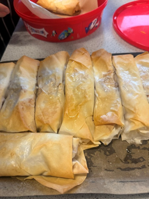 Photograph showing a tray of golden-brown spring rolls arranged in two neat rows. A red container with cartoon characters is partially visible on the left side,