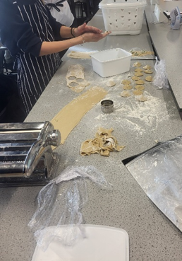 Photograph of a cooking class focused on making pasta, showing students rolling and cutting dough on a flour-dusted countertop. Key elements include a pasta machine, dough cut into circular shapes, and students wearing aprons, highlighting hands-on learning and preparation process.