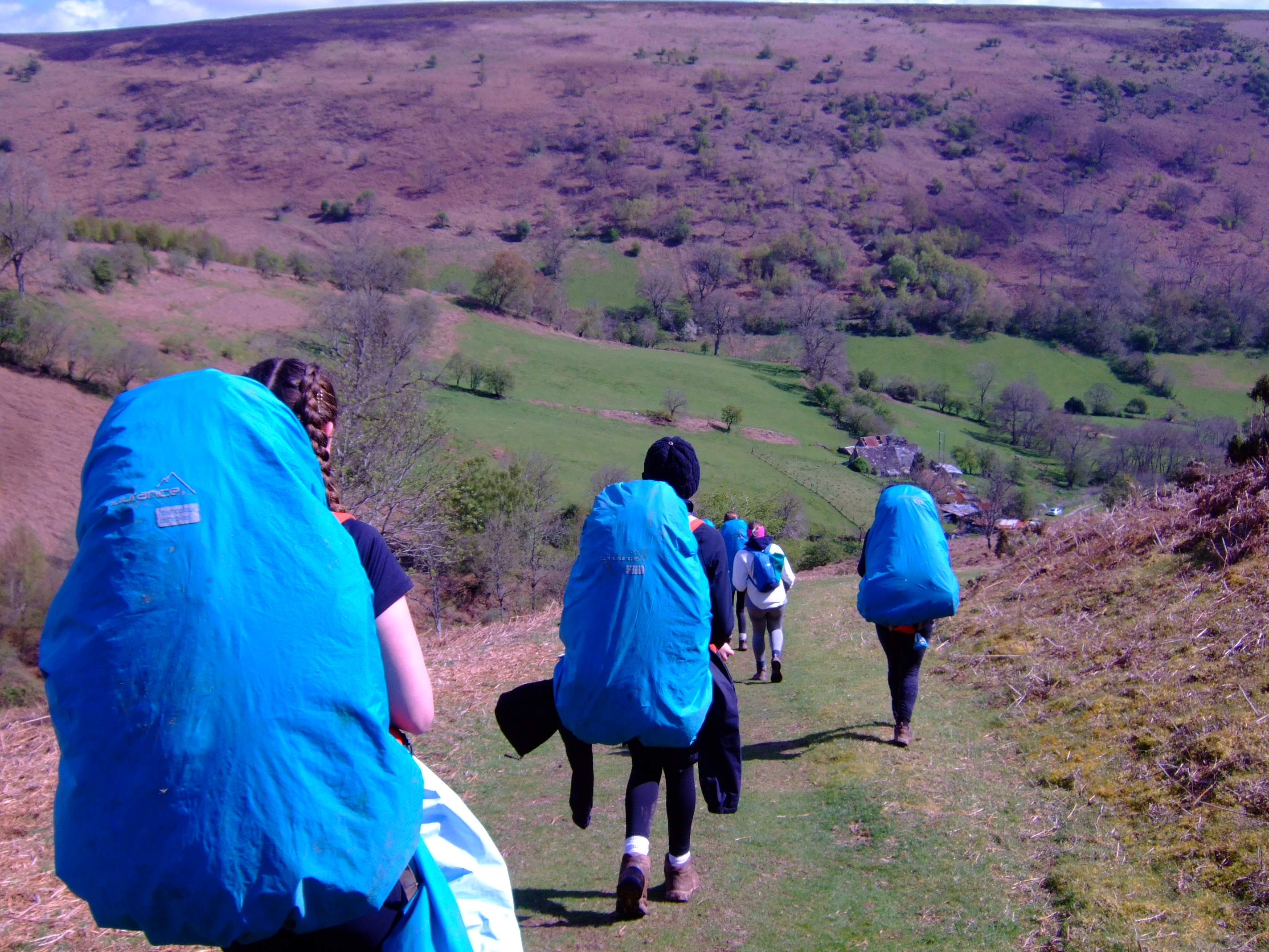 Photograph showing a group of hikers walking down a grassy hillside path towards a valley with scattered trees and a few buildings. Hikers carry large blue backpack
