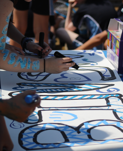 ​​Photograph showing people using black markers to colour and outline large, blue and black patterned letters on a white surface, likely part of a collaborative art or sign-making activity. Visible elements include hands actively drawing, a box of coloured markers, and blue paint or glitter on one person's arm