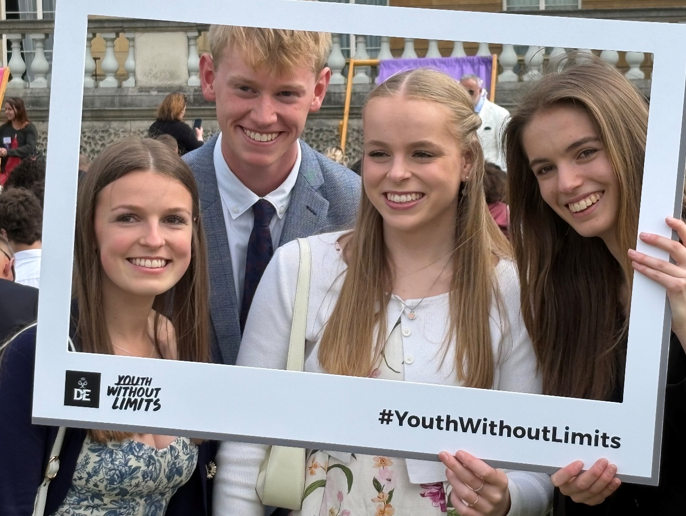 Photograph of four young people posing outdoors while holding a large white rectangular frame with "Youth Without Limits" logo and hashtag #YouthWithoutLimits. Background shows other people and a building with columns, indicating a public or social event.