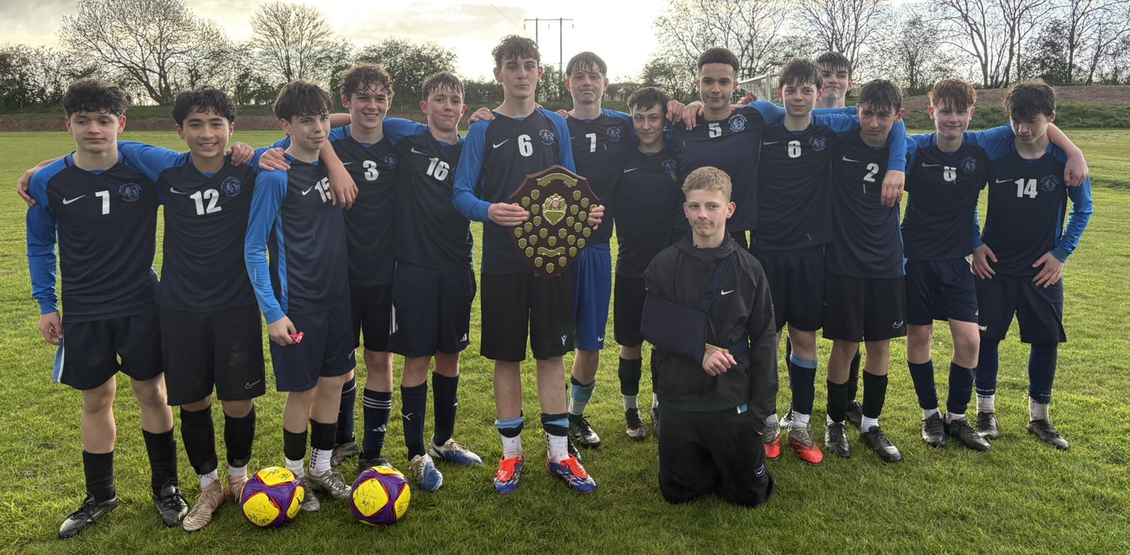 Photograph of a  football team posing outdoors on grass under a cloudy sky, with players wearing black and blue kits. One player holds a large shield-shaped trophy, while two footballs rest on the ground in front