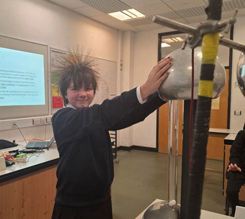 Photograph of a science classroom experiment demonstrating static electricity, where a student’s hair is standing on end after touching a Van de Graaff generator. The scene includes laboratory equipment, a projector screen displaying text, and typical classroom furniture,
