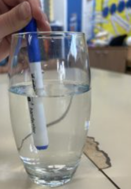 Photograph showing a hand holding a blue and white marker partially submerged in a clear glass of water on a table.