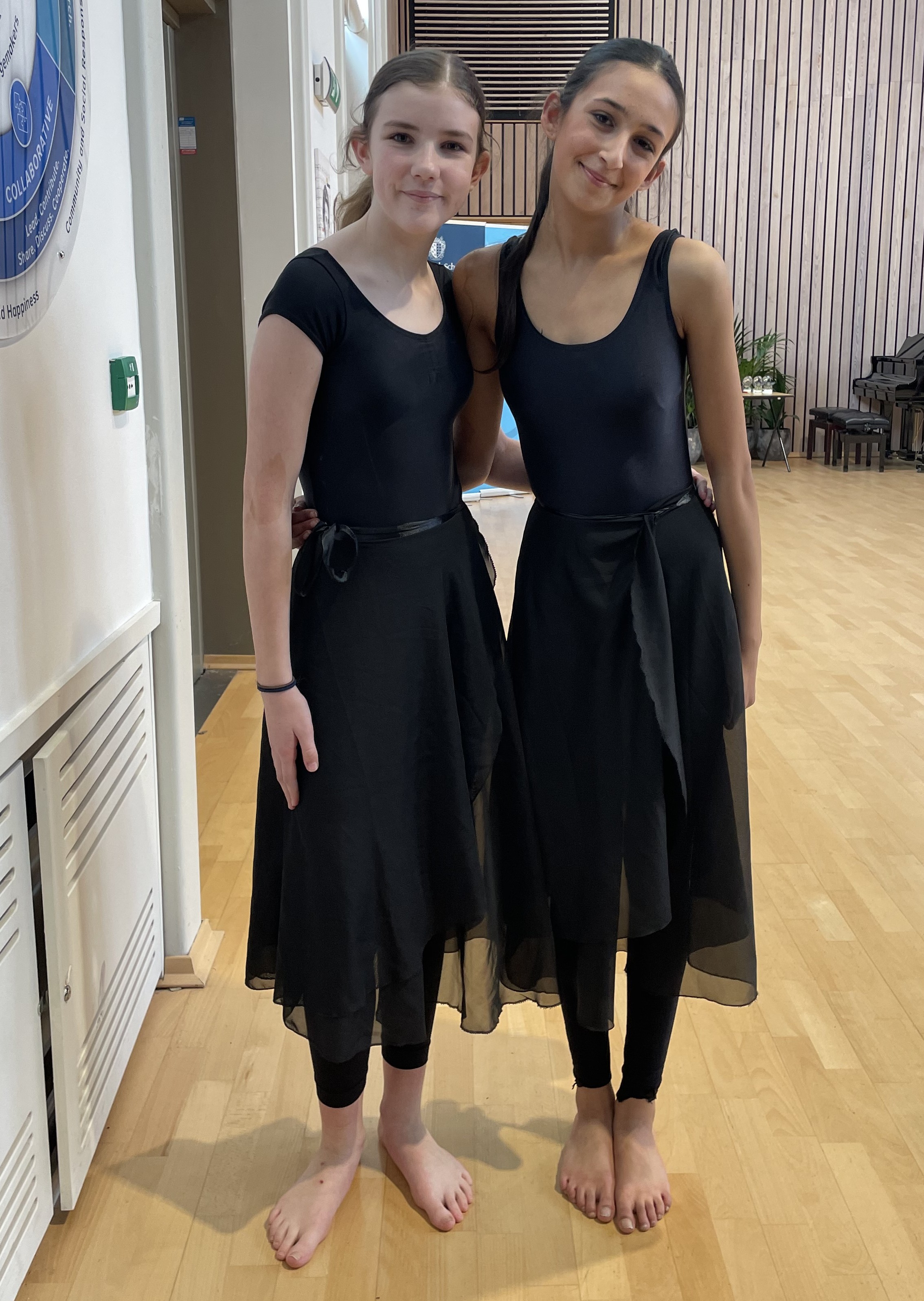 Photograph of two dancers standing barefoot on a wooden floor in a hall. Both wear black leotards and skirts, with a background featuring chairs, a large window, and a wall with vertical wooden panels.