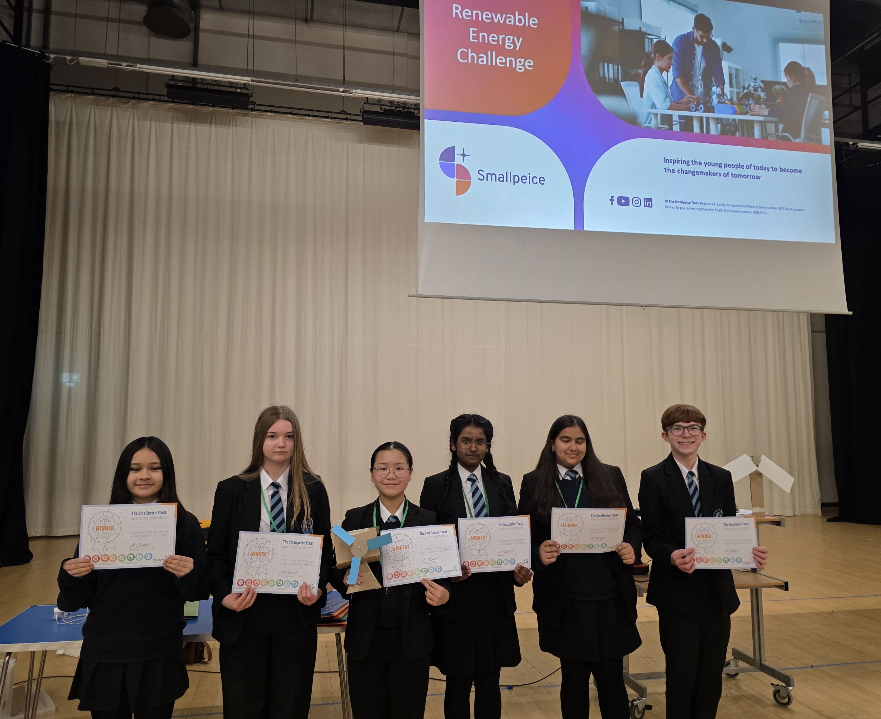 Photograph of six students standing in a row on a stage holding certificates, dressed in school uniform. A large screen behind them displays a presentation slide titled "Renewable Energy Challenge" with Smallpiece logo and supporting text about inspiring young people in STEM
